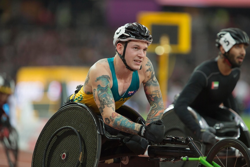 A heavily tattooed man wearing a sleeveless Australian team top steers his wheelchair with his gloved right hand during a race