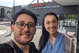 A smiling, dark-haired man takes a selfie with his female partner outside hospital.