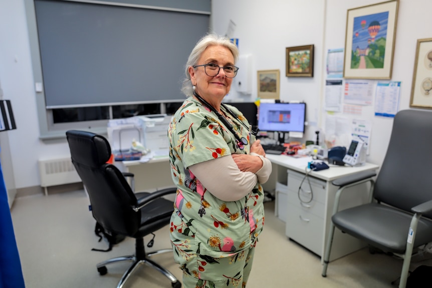 Older woman with white hair and glasses stands side on smiling wearing bright patterned medical scrubs in doctors office