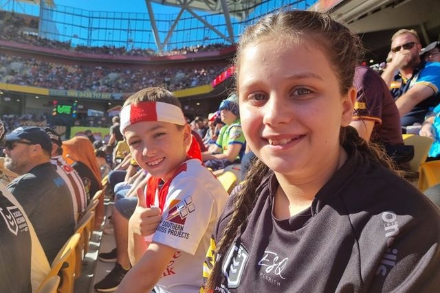 A young girl and boy sitting in a crowd, boy giving a thumbs up, people behind.