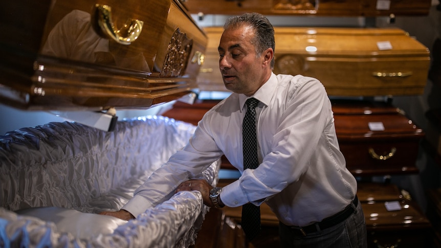 A man wearing a suit examines the satin interior of a coffin on display.