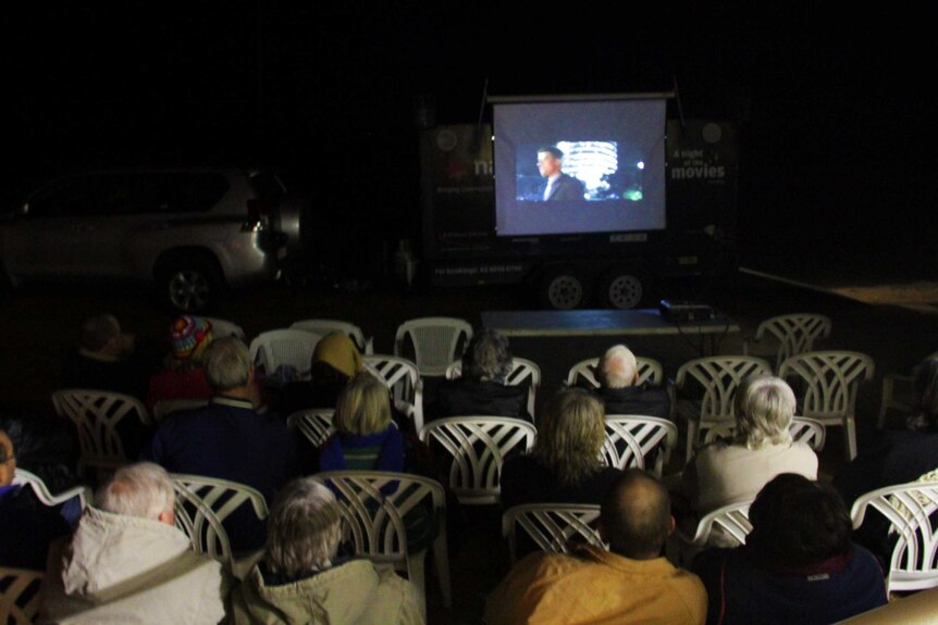 People on white chairs watching a movie on a screen on the back of a trailer.
