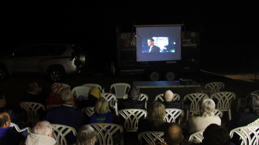 People on white chairs watching a movie on a screen on the back of a trailer.