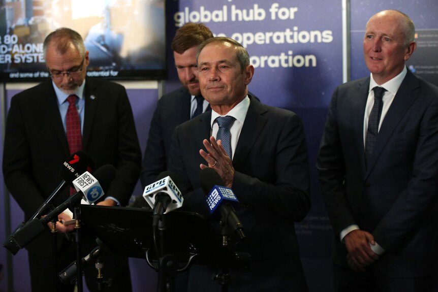 A wide shot of WA Premier Roger Cook speaking at a media conference indoors with three other men standing behind him.