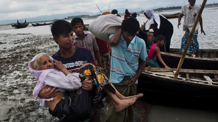 A male Rohingya refugee carries an elderly woman from a small wooden boat.