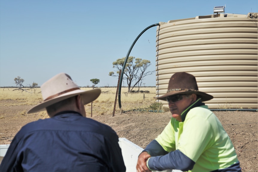 Two men in akubras talk in front of a white water tank with a rectangular device on the roof.