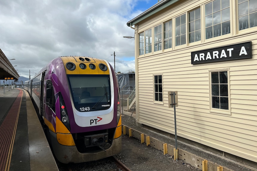 A yellow and purple train at a station.  On the building to the right is a sign saying Ararat A