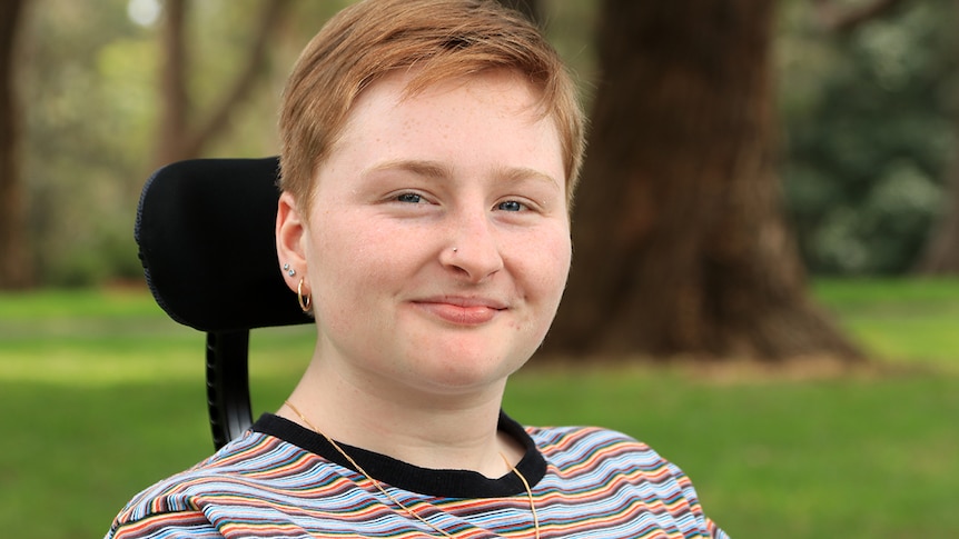 A young woman with short hair looks to the camera and smiles. The black back of her wheelchair is seen against her head.