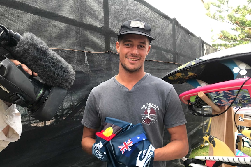 Soli Bailey looks at the camera and holds his wetsuit so the Australian and Aboriginal flags are both visible.