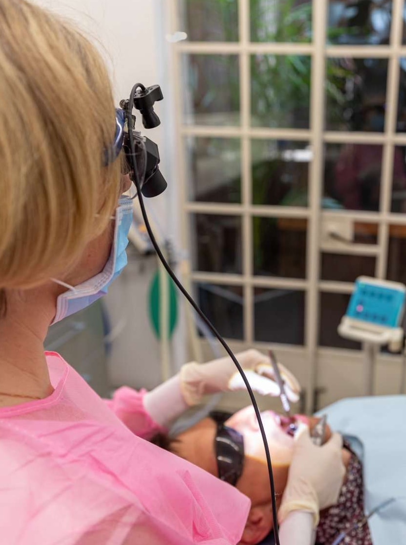 A dentist wearing magnifying eyewear and a paper face mask holds instruments in the mouth of a patient.