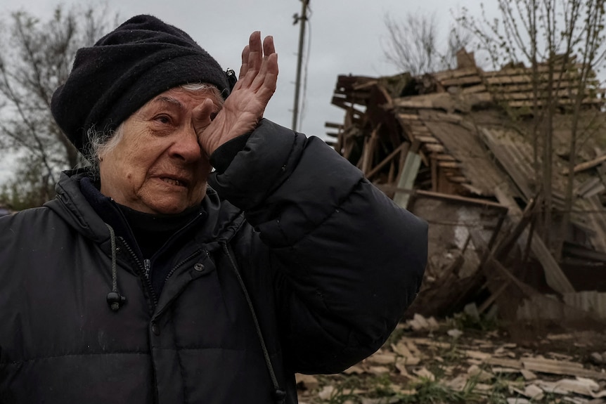 An old lady with grey hair wipes a tear from her eye, looking distressed. She is standing in front of a destroyed structure.