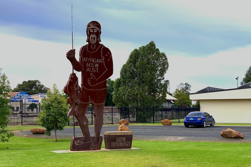 A large metal sculpture of a man in a tank top in thongs holding a fish next to a metal esky.