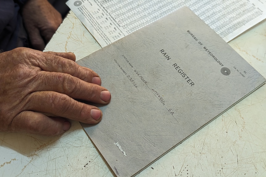 A man's hand at rest on a logbook that says "rain register" on the cover.