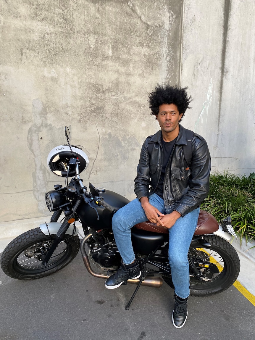 A man with a serious expression in blue jeans and leather jacket, sitting on a motorcycle
