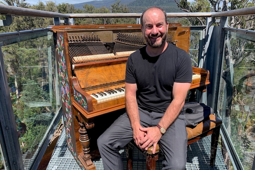 Kelvin Smith sits in front of an upright piano on a cantilevered metal walkway in a forest