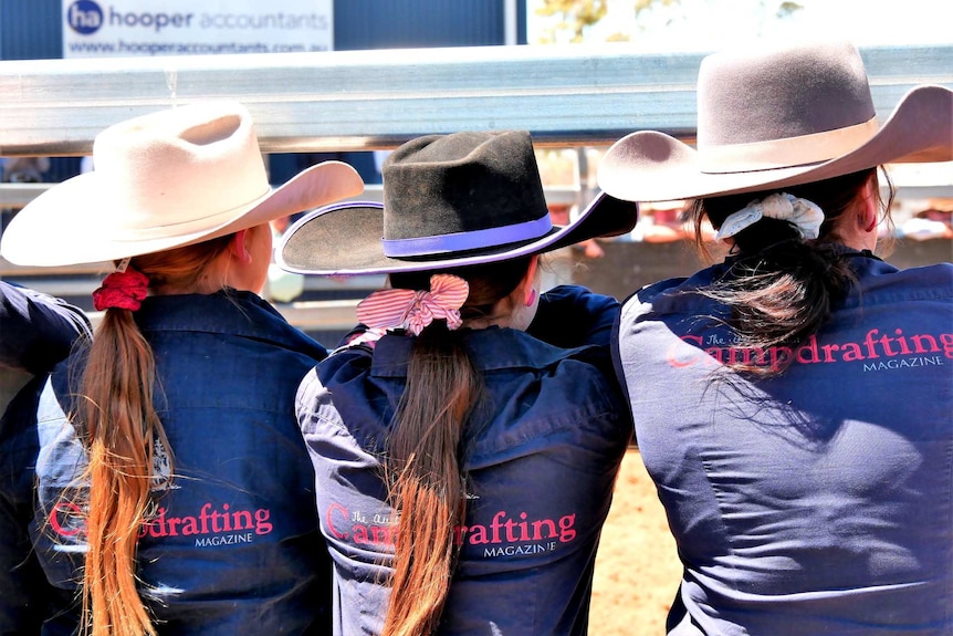 Three girls, watching the campdraft event, wearing purple long-sleeved shirts and hats