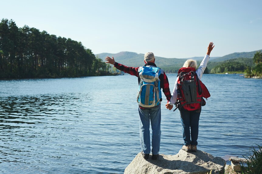 An elderly couple standing on a rock near a lake with their arms up