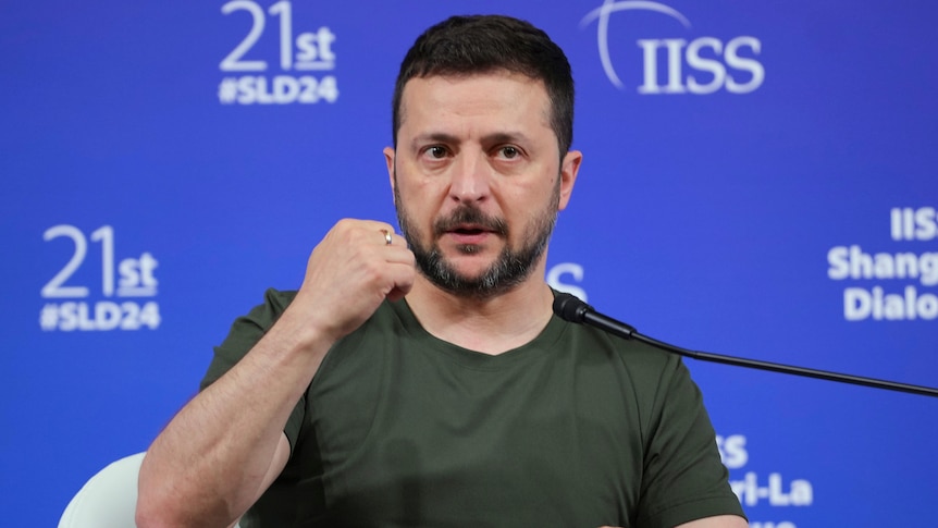 Volodymyr Zelenskyy sits in a dark green shirt holding his fist in the air while making a point at a press conference