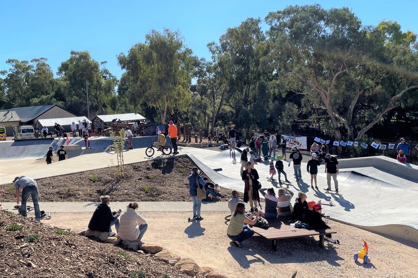 A wide shot of children and youth congregating around a newly built skate park with some bikes and boards on ramps.