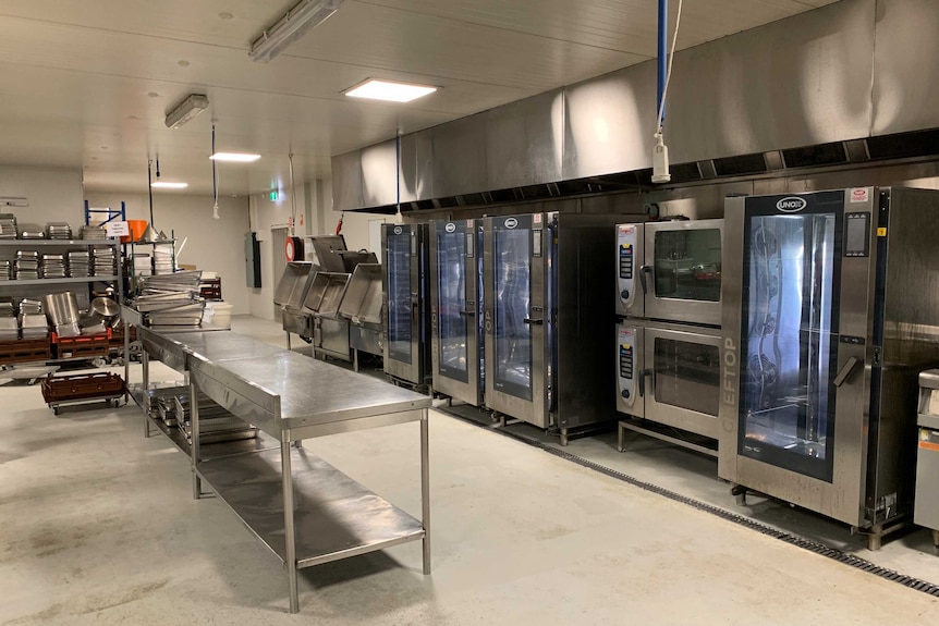 A commercial kitchen, with stainless steel benches, industrial fridges and neat stacks of baking trays.