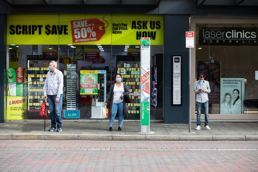 Three people social distancing while wearing masks at a bus stop.