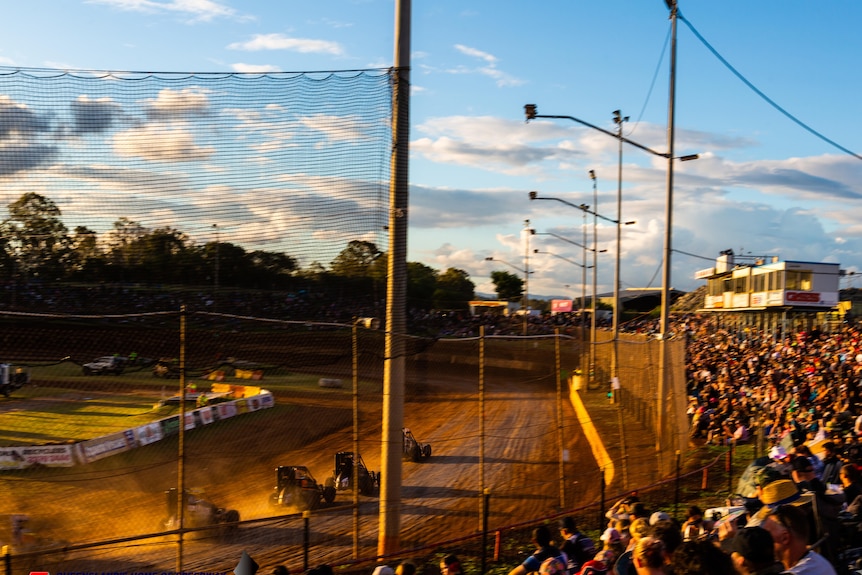 Speedcars kick up dirt along a track as pack crowds look on from the side.