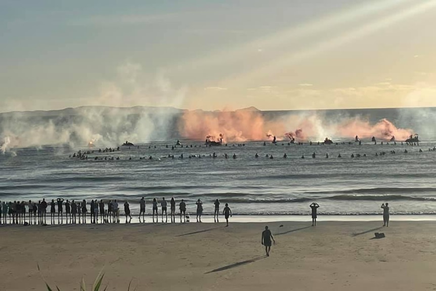 Coloured smoke billows over the water where a group of surfers on boards watch.