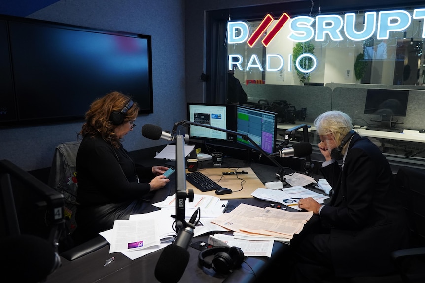 Libbi and Bob sitting in a radio studio in front of microphones, D//srupt Radio sign in window.