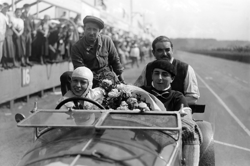 In this black and white photo, two racing drivers are pictured at Le Mans, with two passengers sitting at the back of the car.