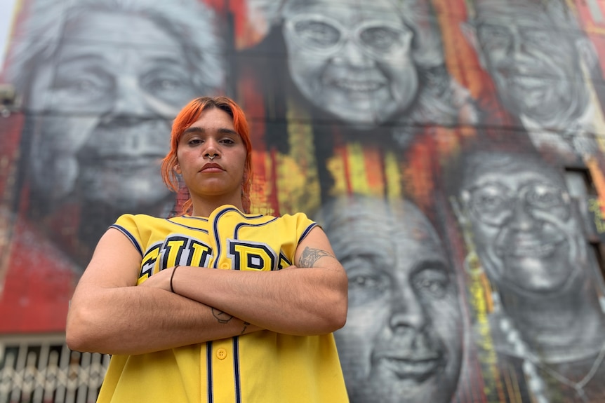 Sky Thomas stands with her arms crossed, in front of a street art mural in red, yellow and black.