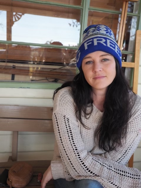 Woman with dark hair and a blue beanie on looks at the camera. She is sitting outside and is wearing a woolen jumper.