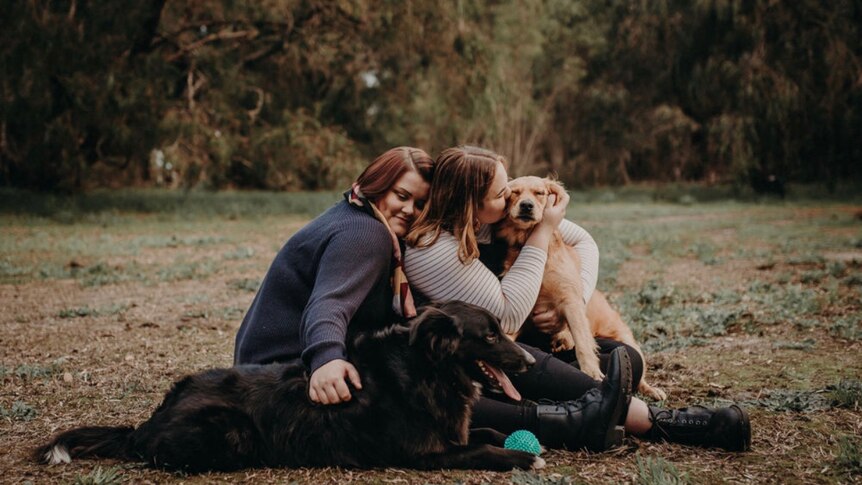Two women sit in a field, hugging their black and brown dogs.