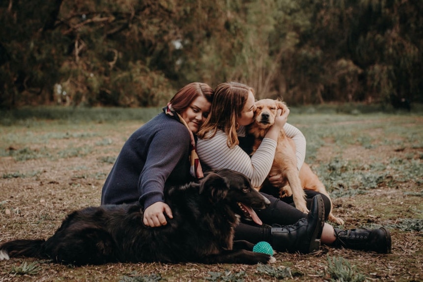 Two women sit in a field, hugging their black and brown dogs.