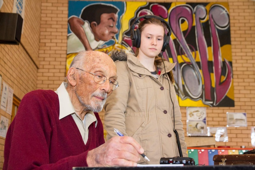 A man with recording equipment sitting down and a teenaged girl standing with headphones on.