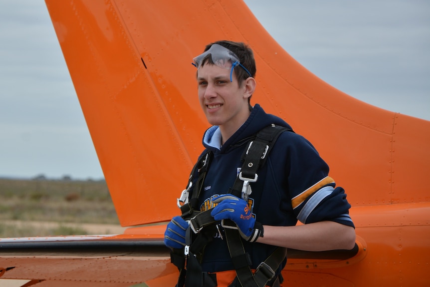A boy in skydiving equipment stands next to a plane.