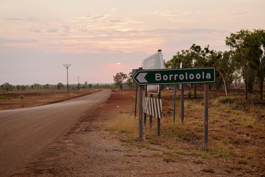 A road sign saying Borroloola next to a road at sunset