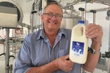 John Cochrane smiles as he holds up a bottle of milk with a vat behind him.