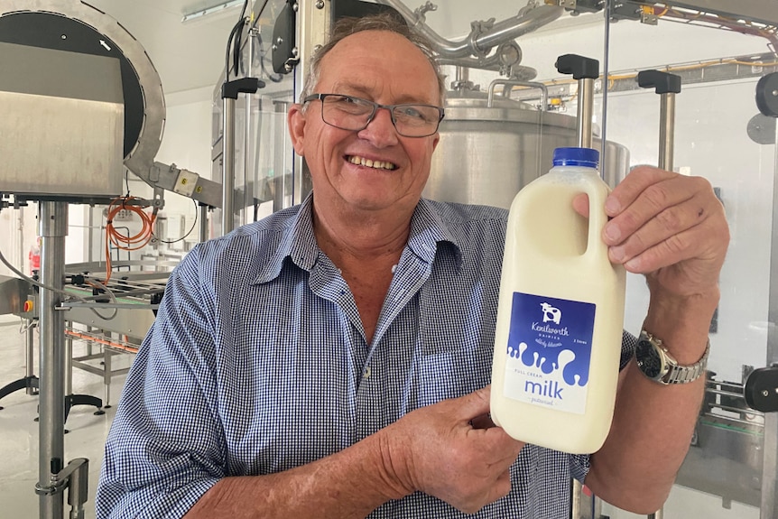 John Cochrane smiles as he holds up a bottle of milk with a vat behind him.