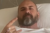 A man in a hospital bed, with a canula in his hand, does a hang loose symbol. 