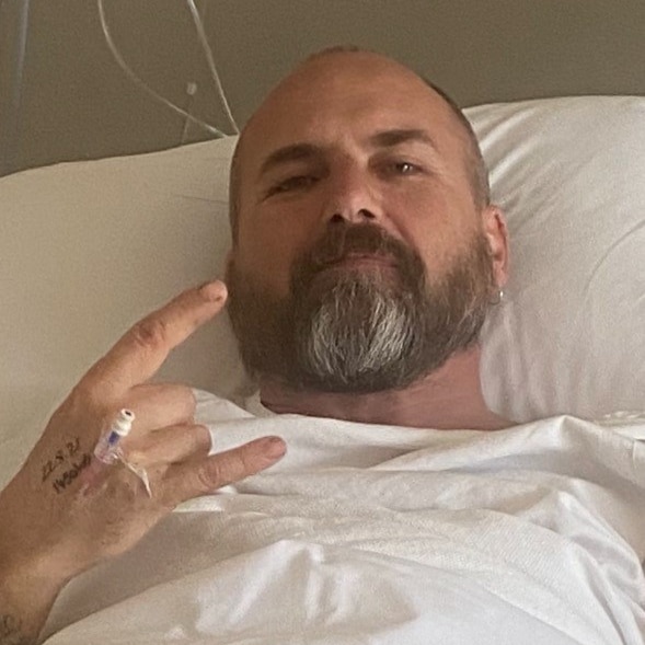 A man in a hospital bed, with a canula in his hand, does a hang loose symbol. 