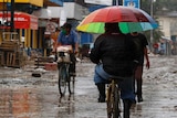 Man rides a bike with a rainbow umbrella on a muddy and messy street.