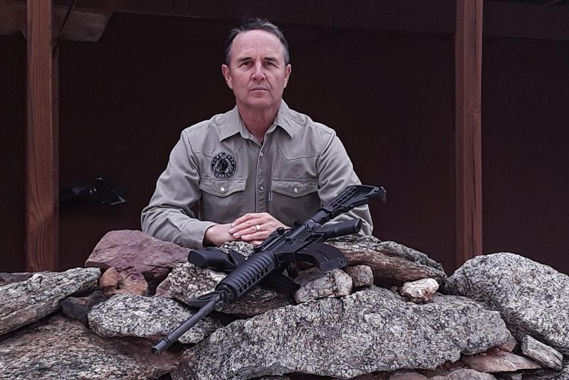 Drew Miller sits with his hands folded staring at the camera with a gun in front of him.