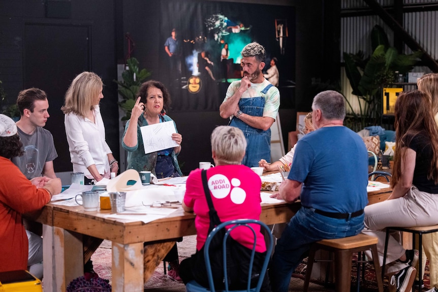 A group of ten people stand around a large wooden table. Wendy gestures to a paper scribbled on.