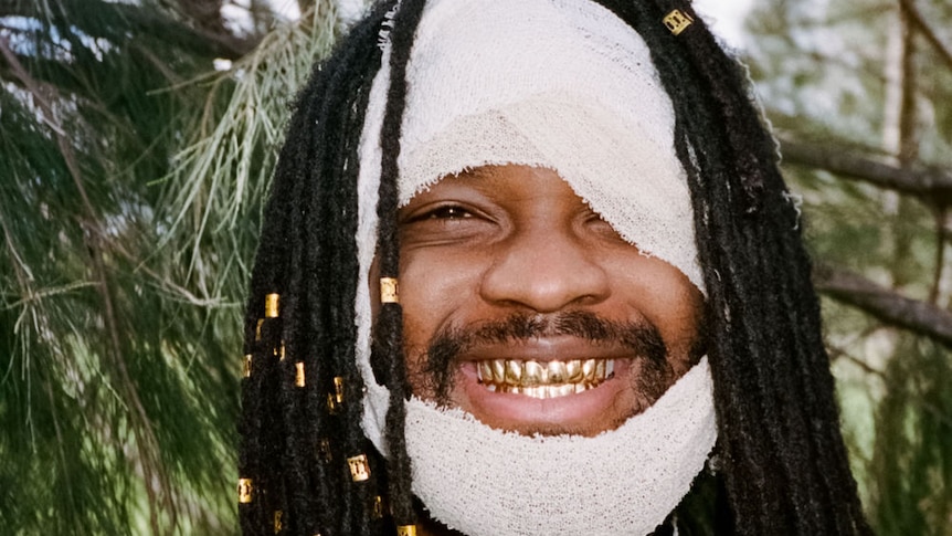 A man is smiling with gold grills, his face is covered in a white bandage, with only his right eye, nose and mouth visible.