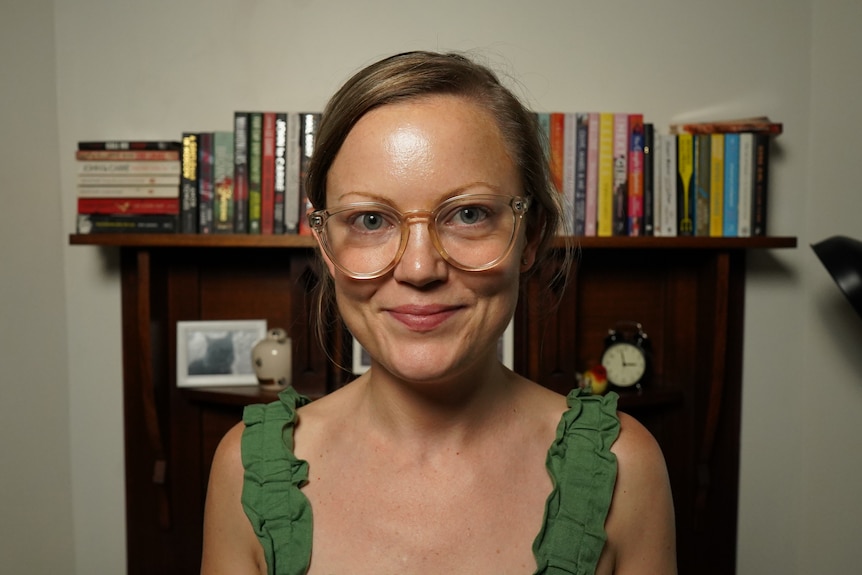 Young woman with glasses in a green top standing in front of a desk with books along the shelf.