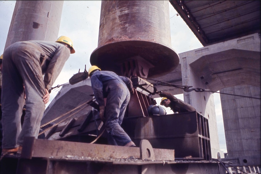 Men in grey overalls and yellow hats work on a large construction