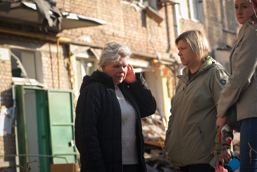 A middle-aged woman talks to two other women in front of a badly damaged building.