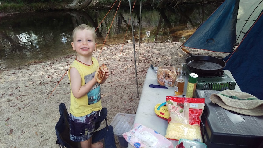 A photo of toddler Sonny standing in front of a tent eating a sausage on bread on a camping trip