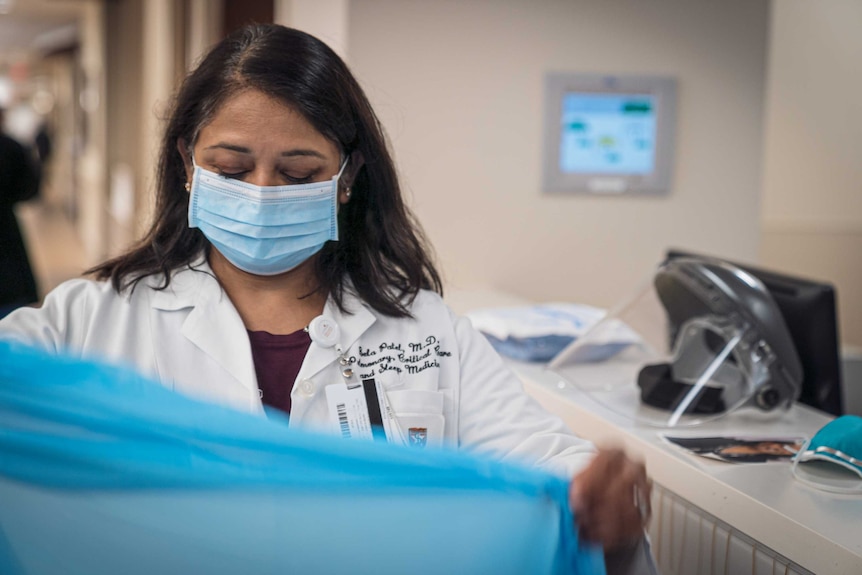 A doctor in a lab coat and face mask opening a blue gown