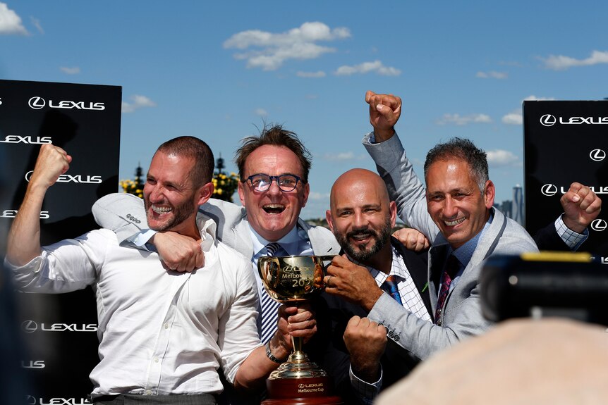 A group of cheering owners stand holding the Melbourne Cup trophy